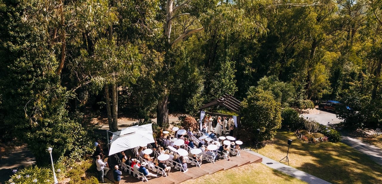 The garden ceremony from above, guests on the timber deck under market umbrellas among tall gums