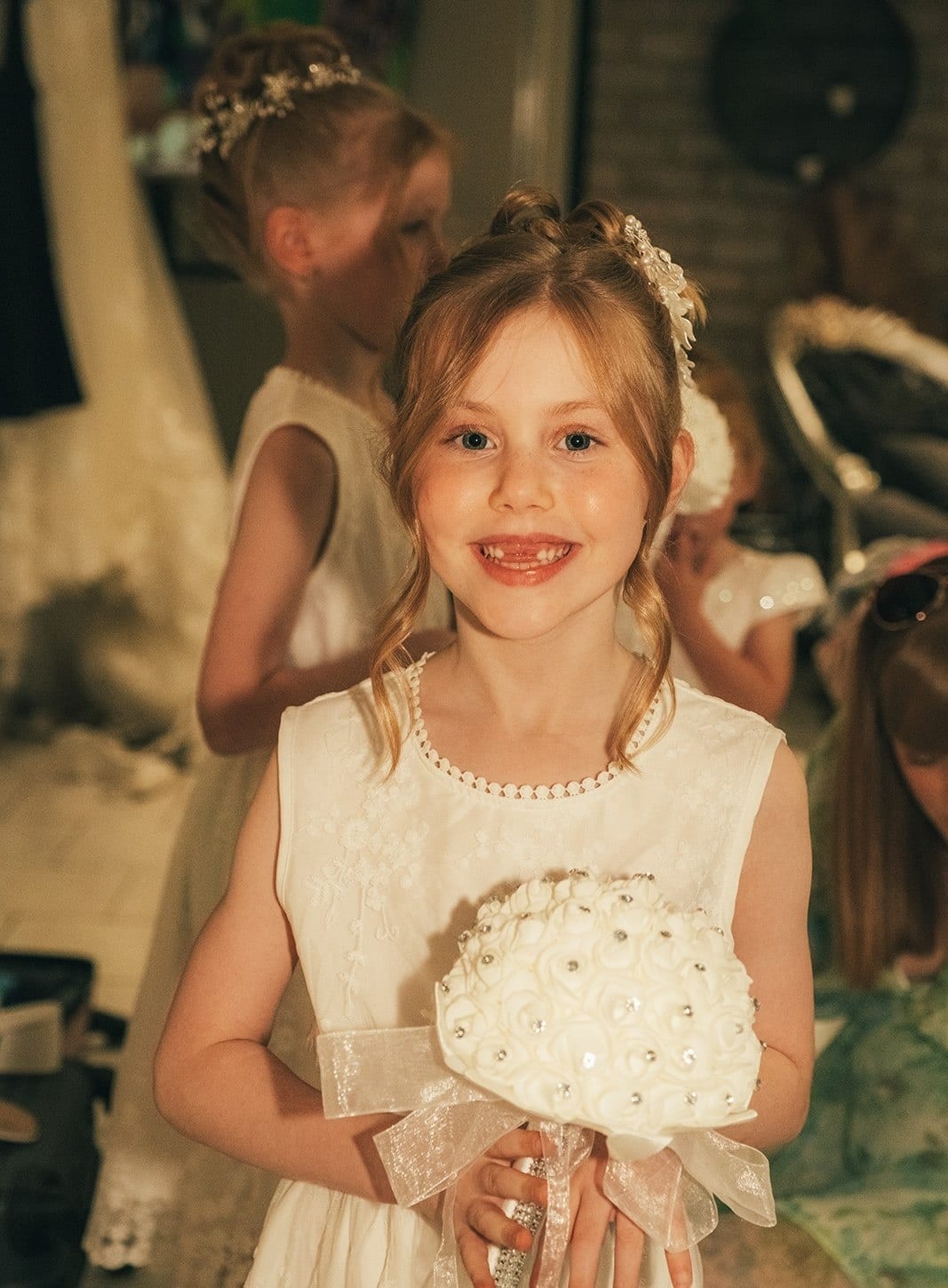 A flower girl in a white dress smiling and holding a round white bouquet