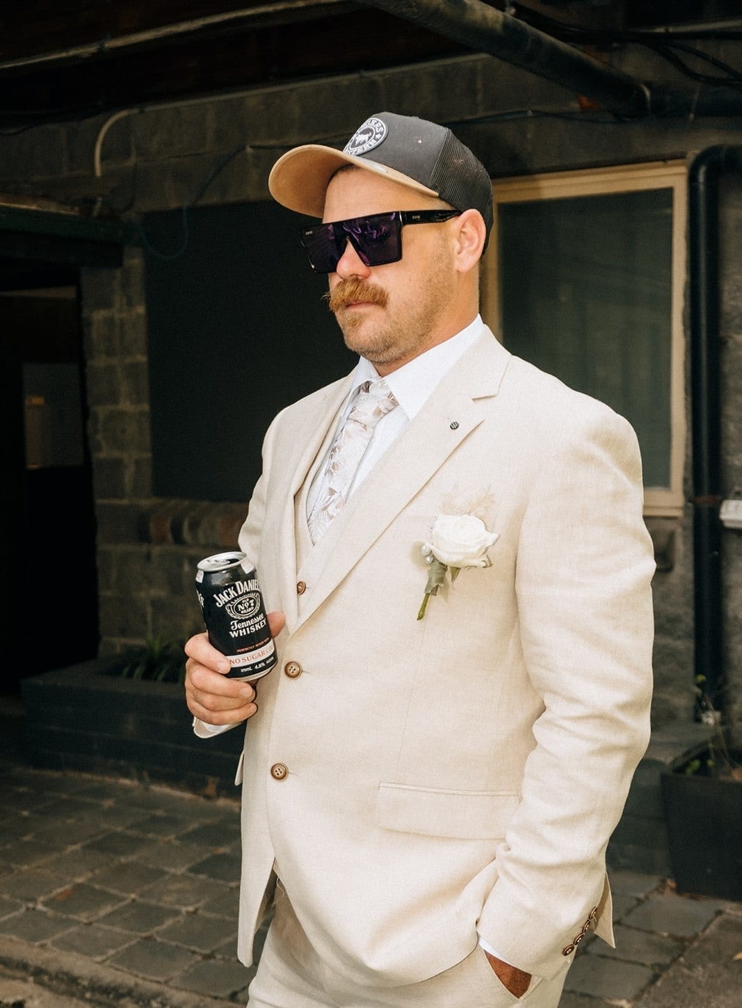 A groomsman in a cream suit, cap and sunglasses holding a drink by the door