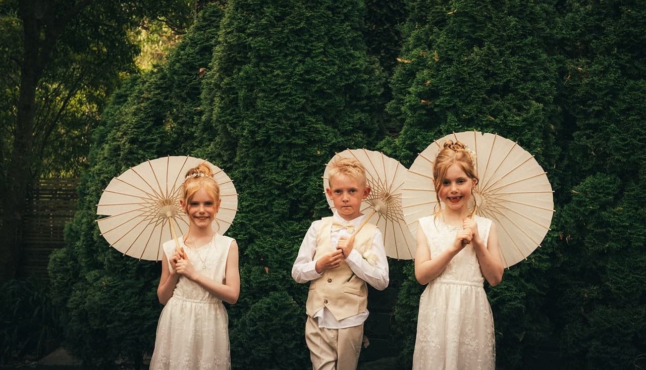 Three children holding white parasols in front of a dark green hedge