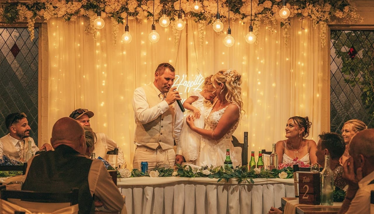 Nick speaking into the microphone at the bridal table, Jenna smiling beside him