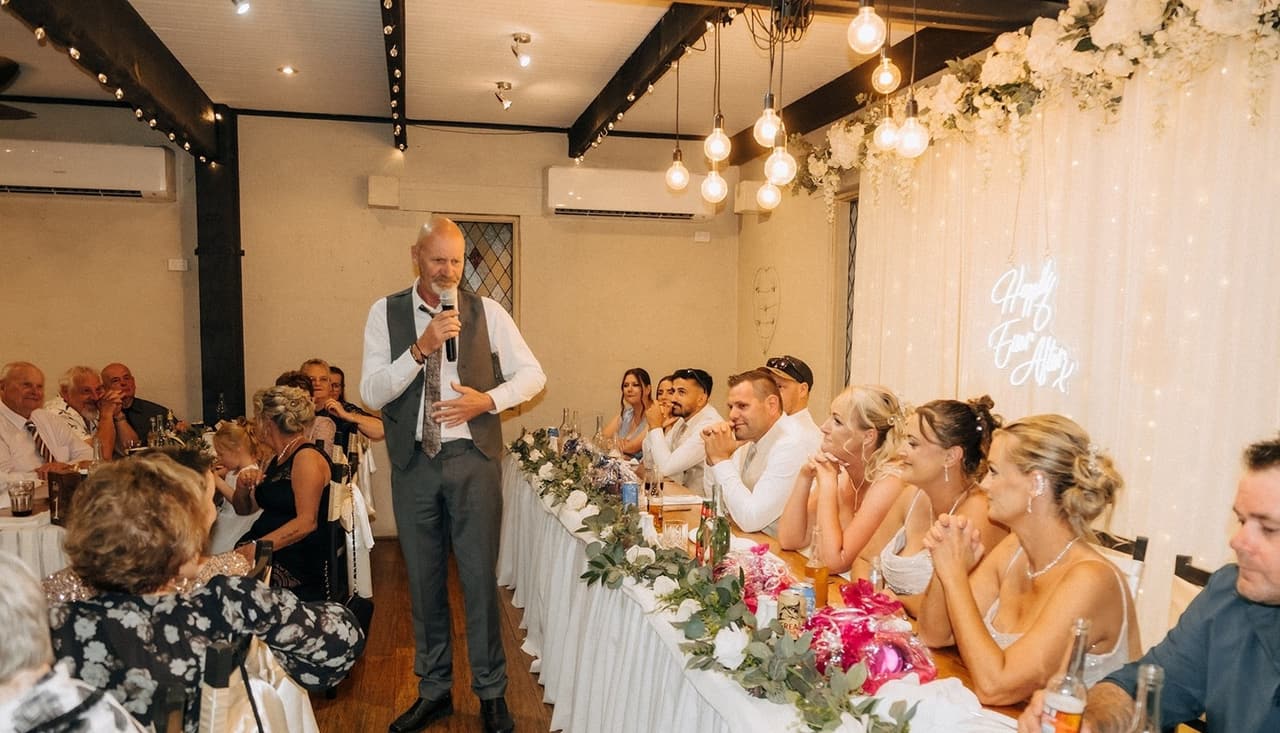 A guest giving a speech in front of the bridal table, the bridesmaids laughing