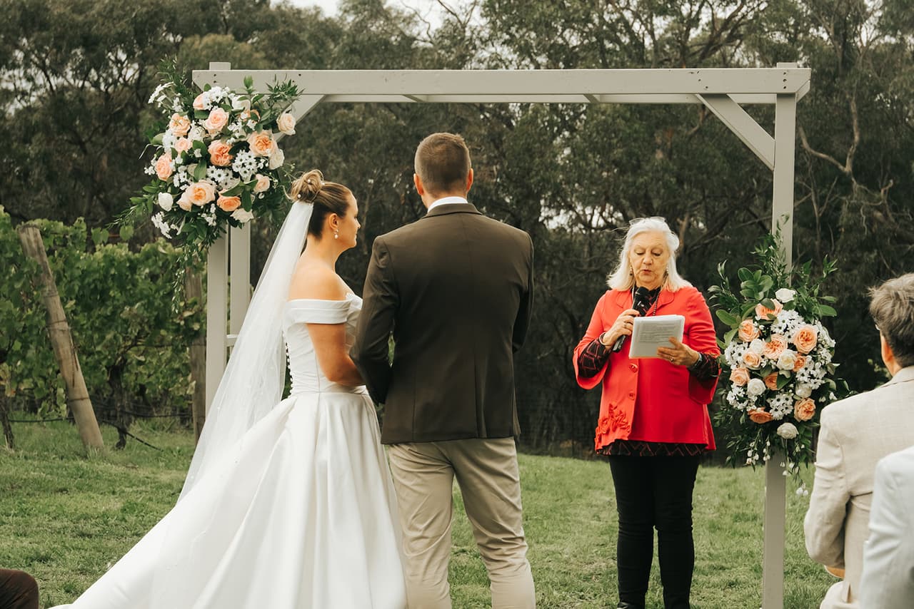 Jessica and Max at the white arbour with its pink and peach flowers, the celebrant in red reading beside them