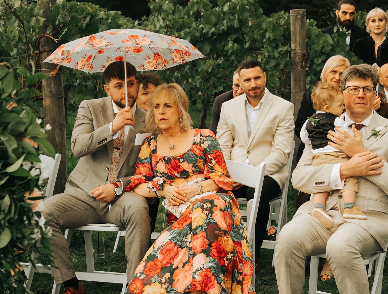 A young man holding a floral umbrella over a woman in a bright floral dress as they watch the ceremony