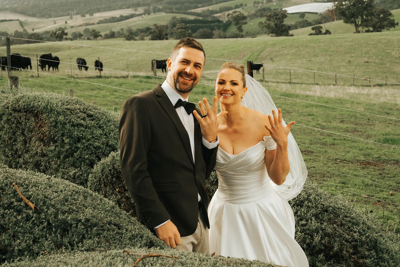 Jessica and Max holding up their rings among the rounded hedges, cattle grazing in the paddock behind
