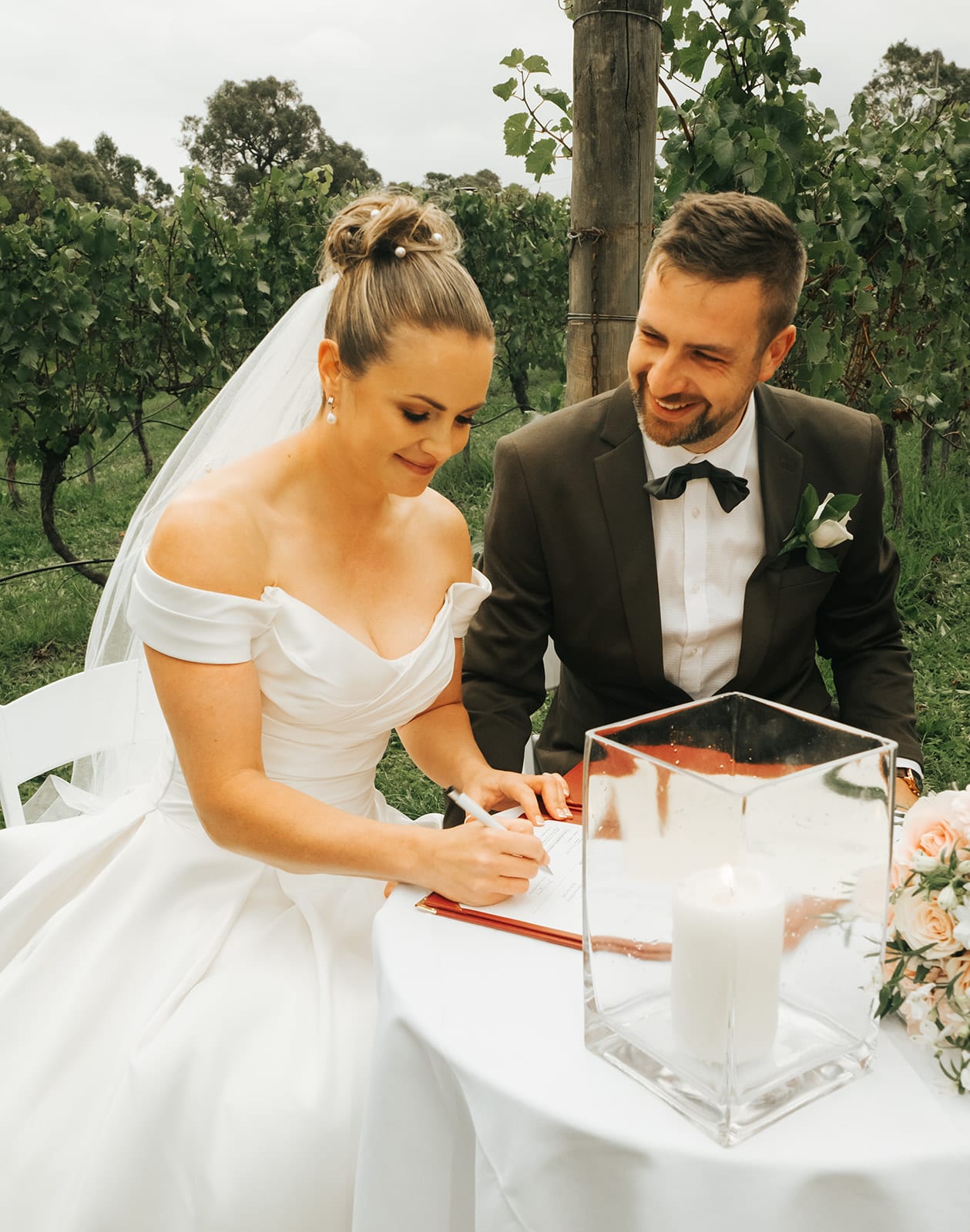 Jessica signing at a round table among the vines while Max smiles beside her