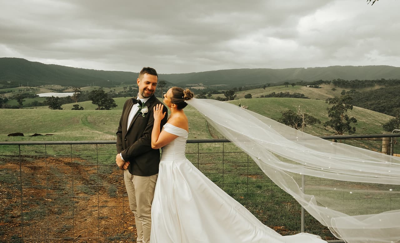 Jessica’s veil streaming out over the paddock fence as Max smiles, hills behind them