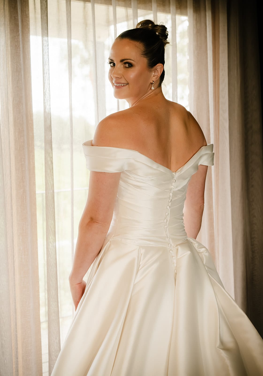 Jessica looking back over her shoulder in her off-shoulder gown beside a sheer-curtained window