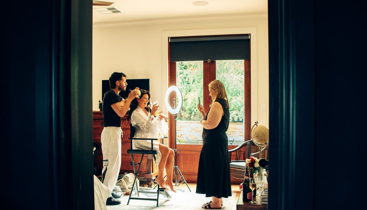 Joanna having her hair done in a sunlit bedroom, seen through a doorway framed by dark curtains