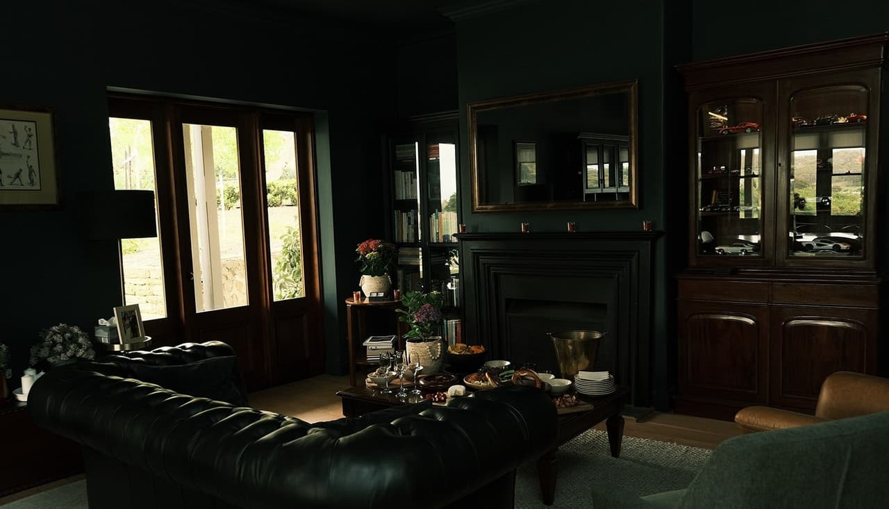 The dark lounge room of the house, with a leather sofa, a fireplace and tall windows