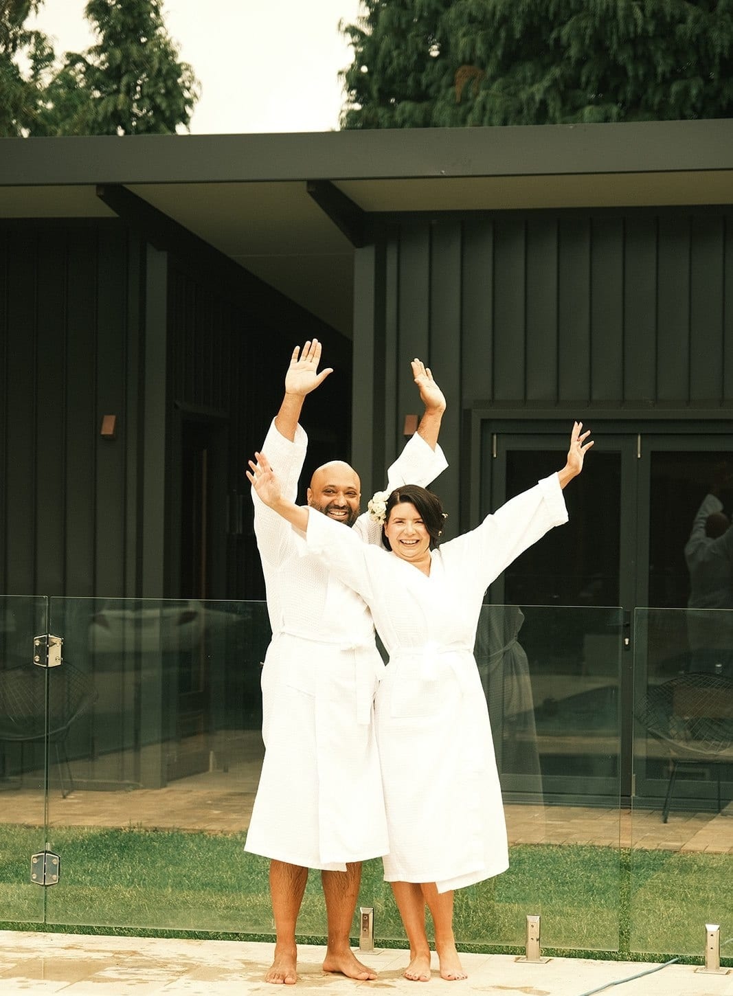 Joanna and Krish in white robes by the pool with their arms in the air