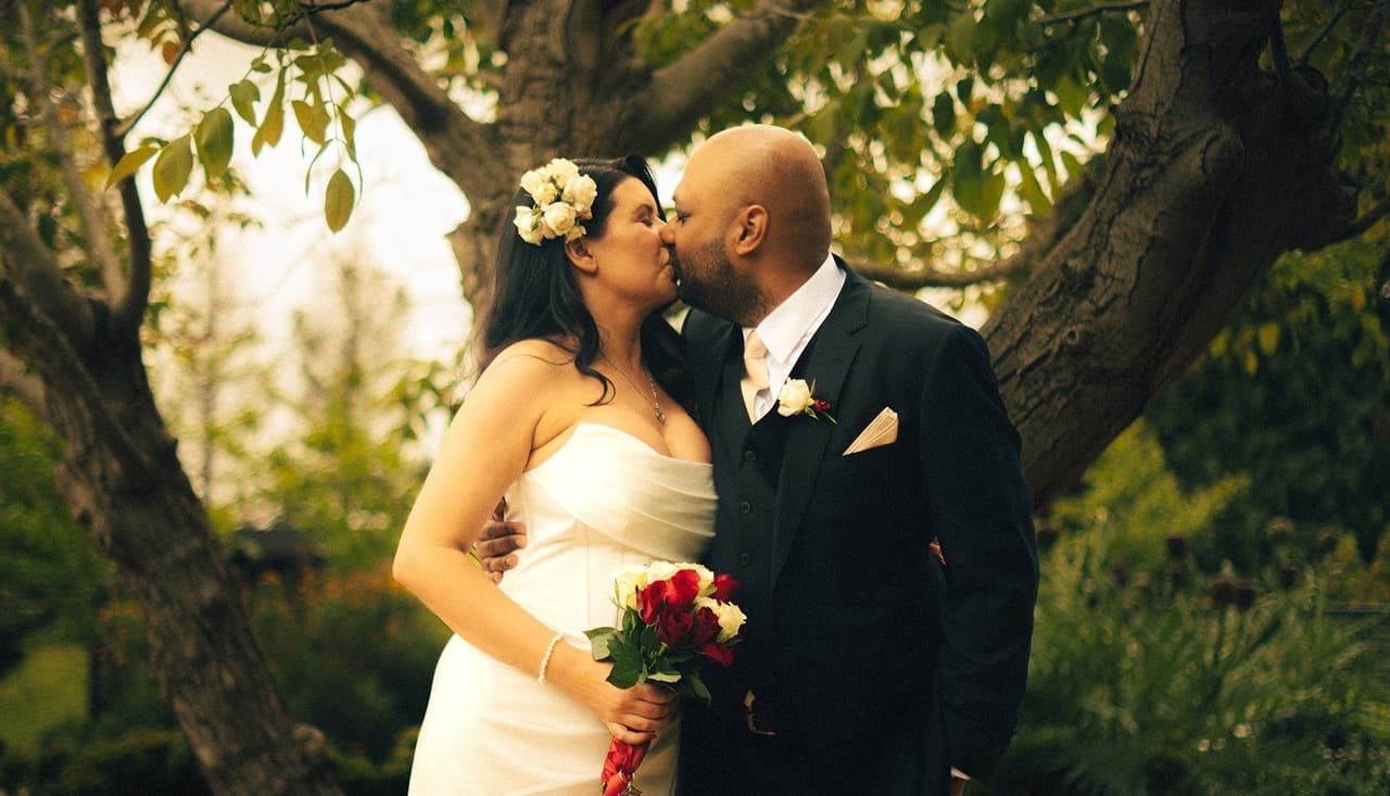Krish kissing Joanna on the cheek under the tree, her bouquet in hand