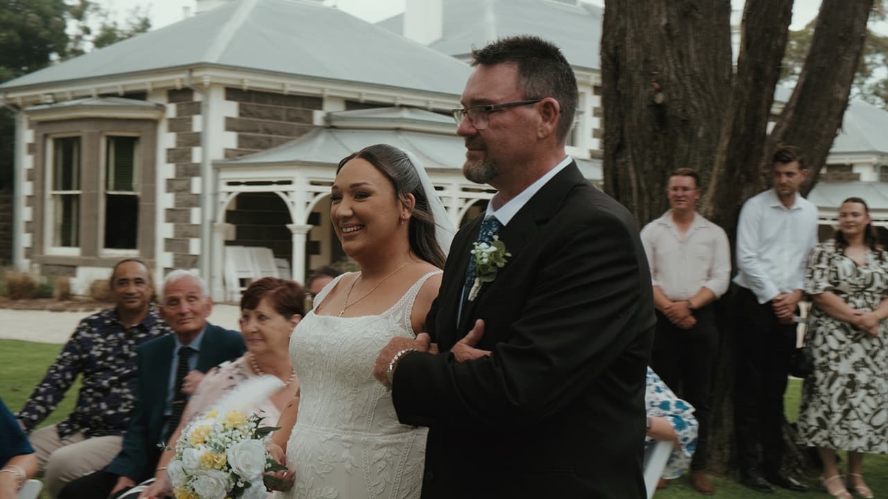 Karina smiling on her dad’s arm as they walk down the aisle, the homestead behind them