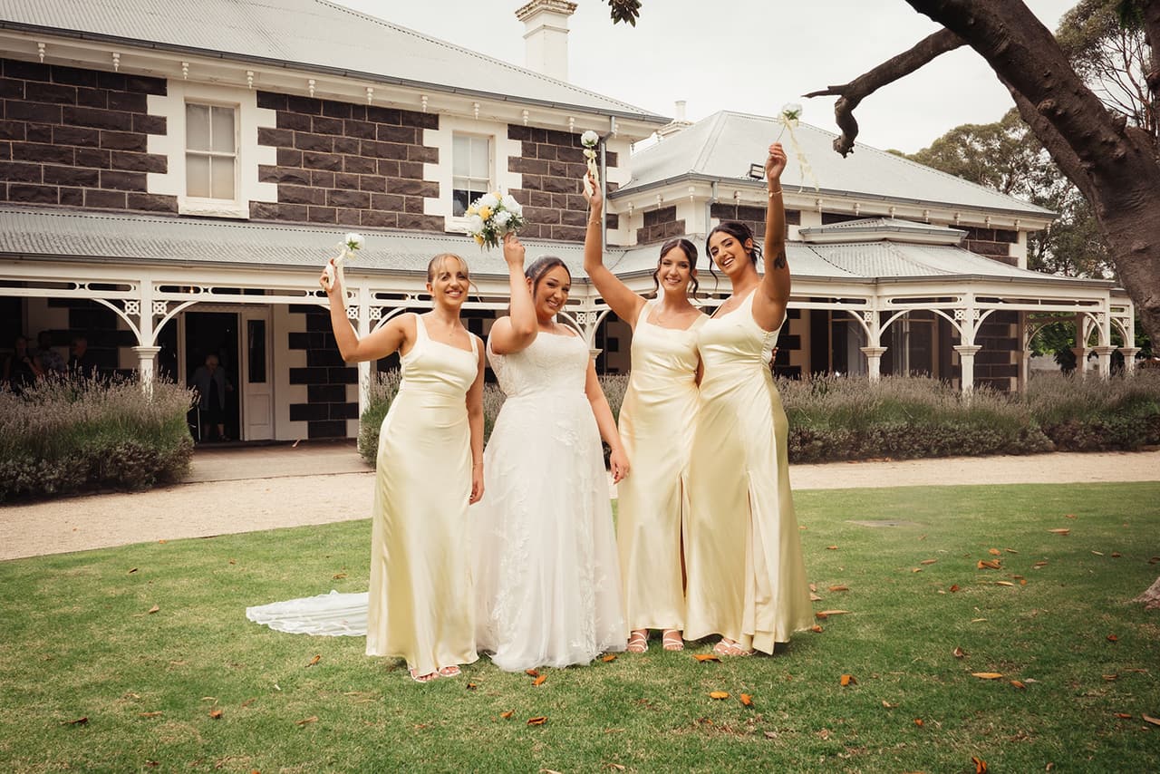 Three bridesmaids in champagne gowns cheering with their bouquets in front of the bluestone homestead