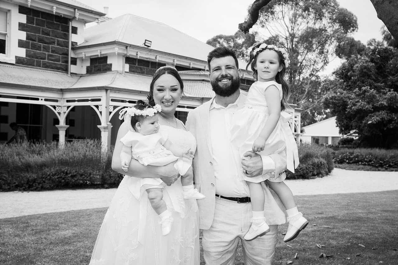 Black and white photo of Karina holding a baby and Bronson holding the flower girl in front of the homestead