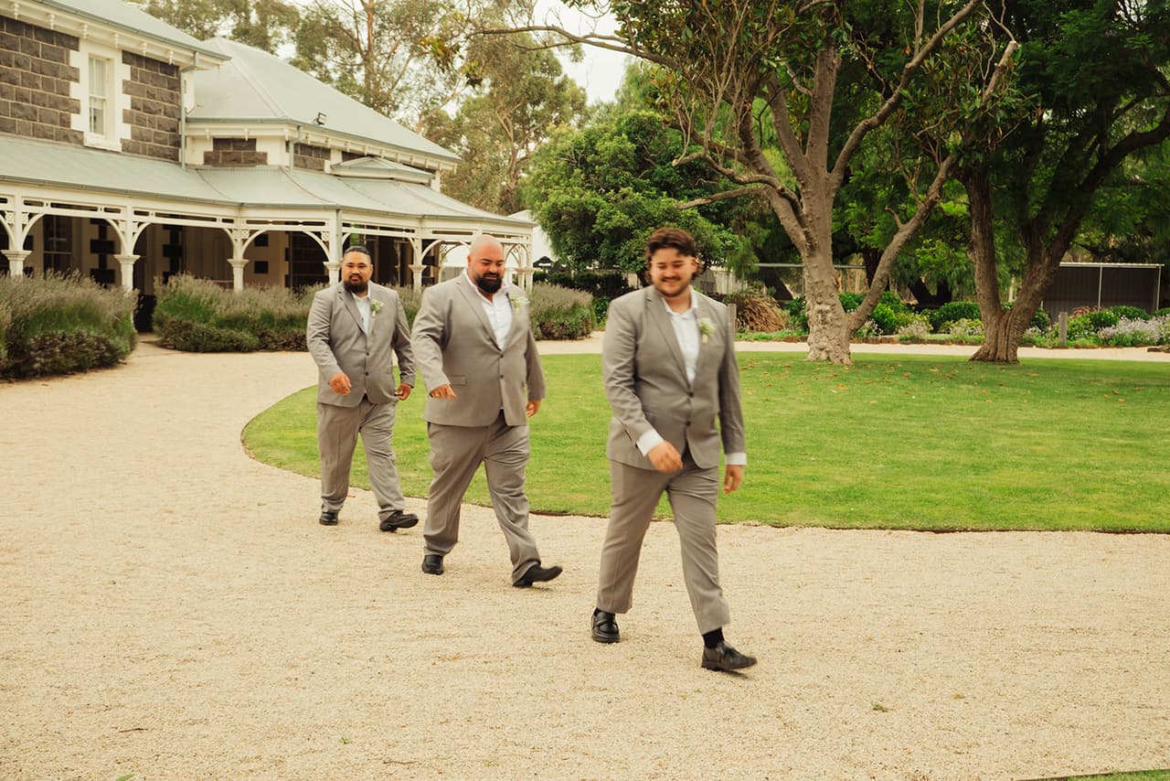 Three groomsmen in grey suits walking along the gravel drive past the homestead