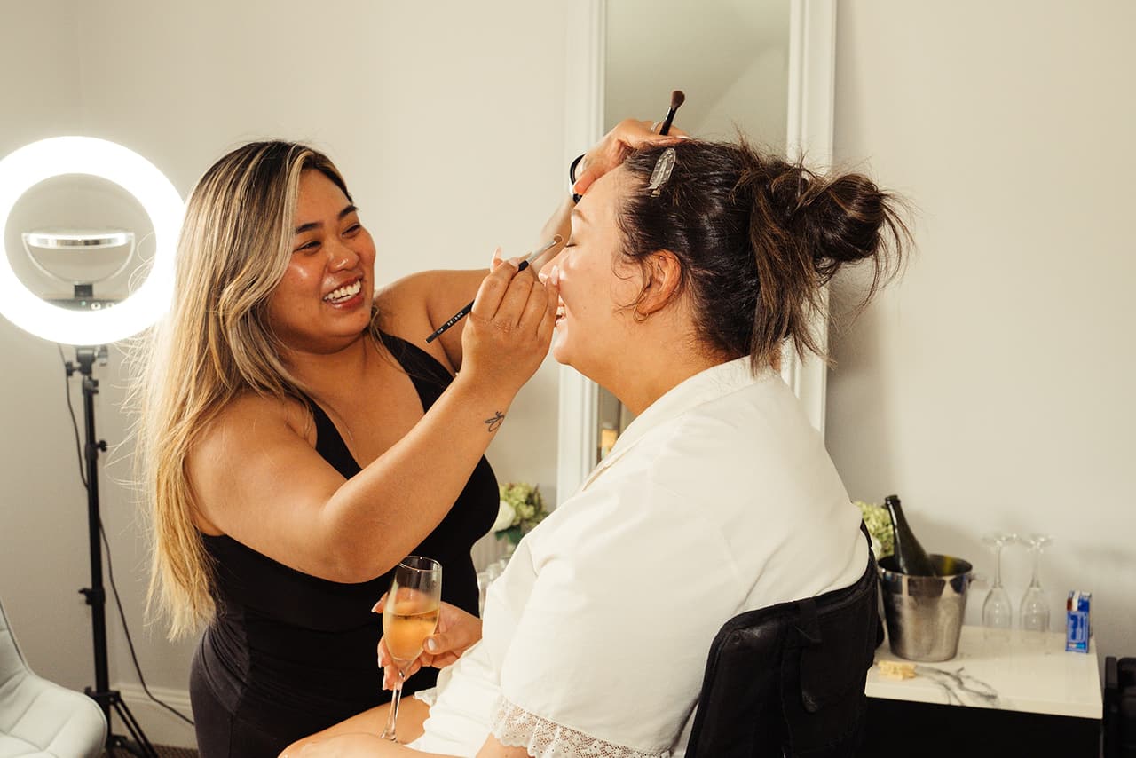The makeup artist laughing as she works on Karina’s makeup beside a ring light
