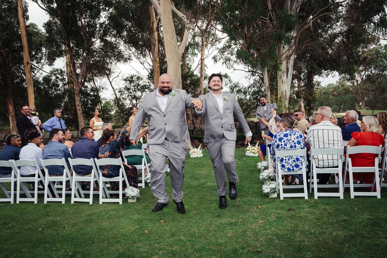 Two groomsmen in grey suits striding down the grass aisle past the seated guests