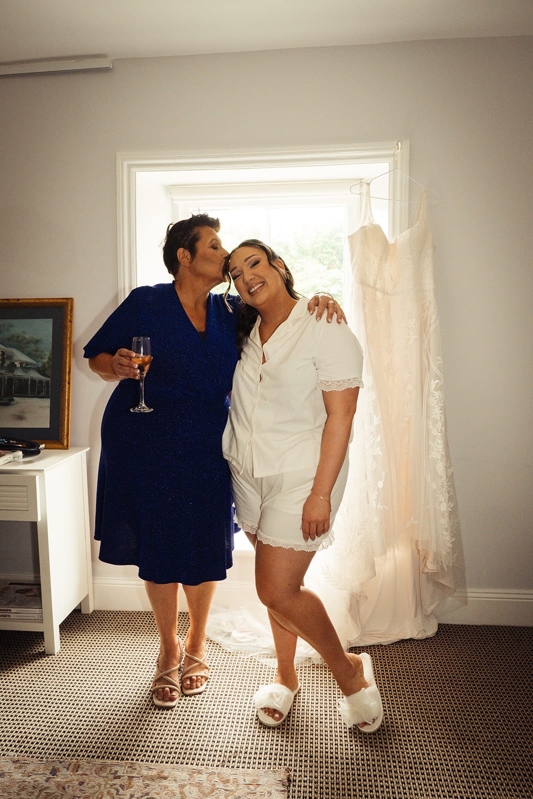 A woman in royal blue kissing Karina’s head by the tall window, the gown hanging behind them