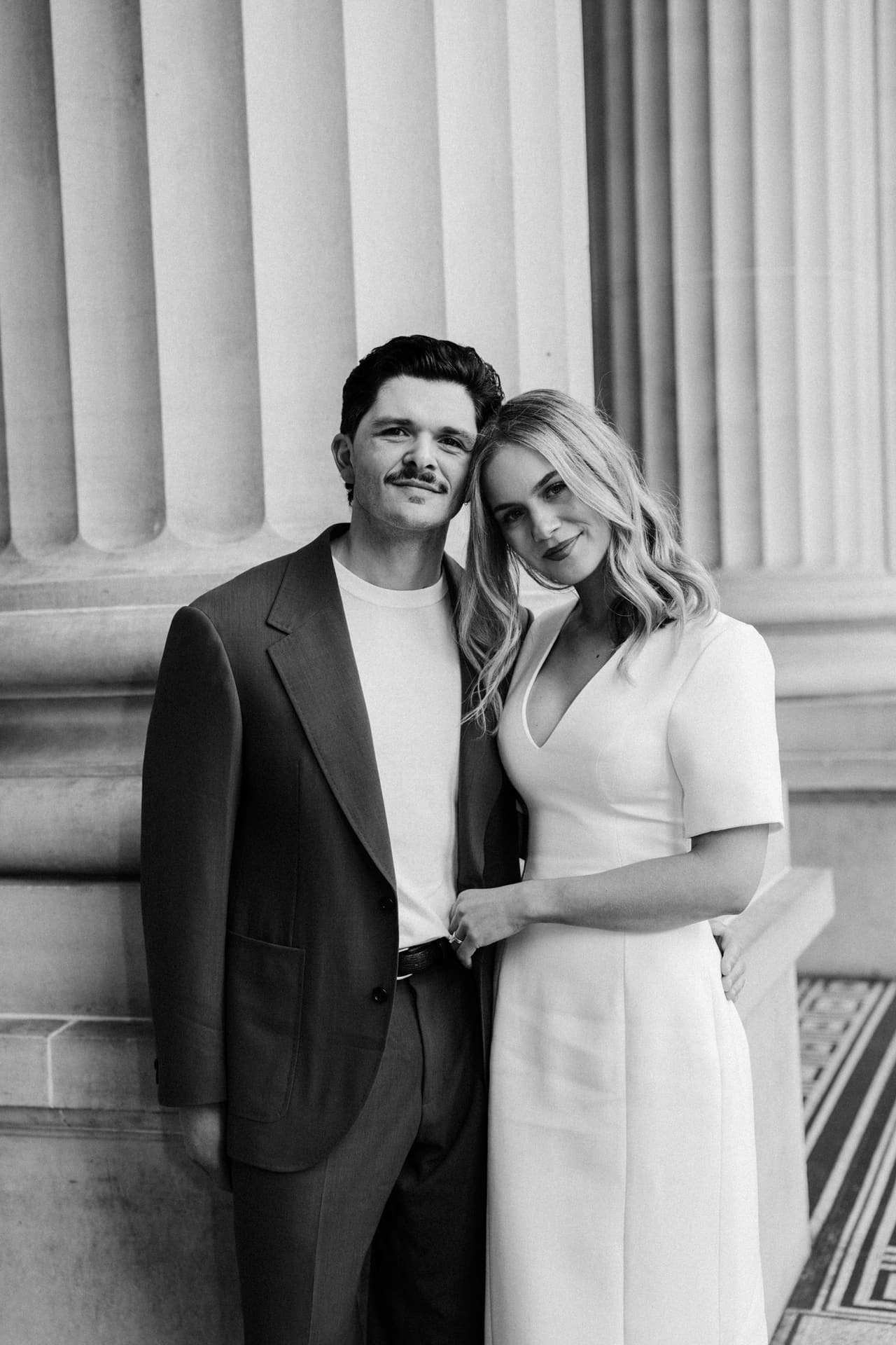 Black and white portrait of Karina and Chris standing close in front of the columns of Parliament House