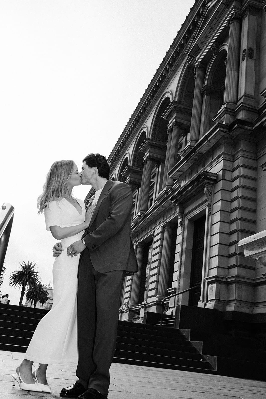 Black and white photo of Karina and Chris kissing beside the arched windows of the Old Treasury Building