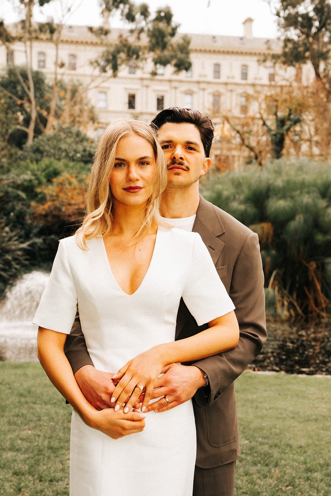 Chris holding Karina from behind on the lawn, their hands and rings together, a fountain behind them