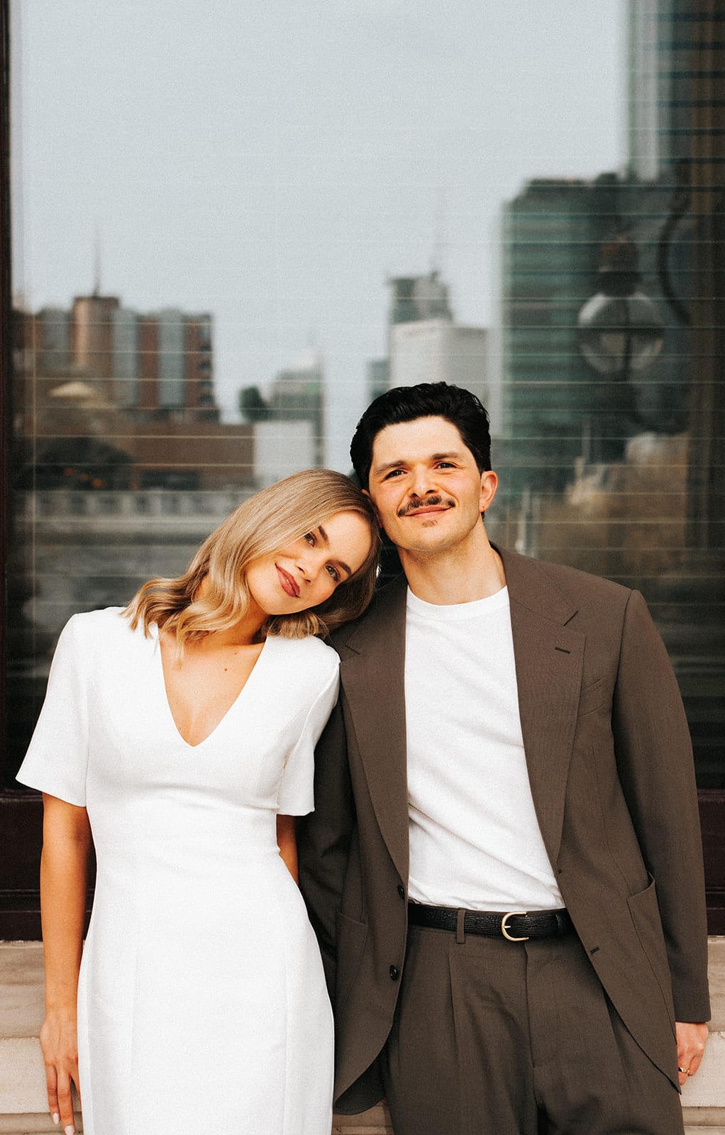 Karina resting her head on Chris’s shoulder in front of a tall window that reflects the city
