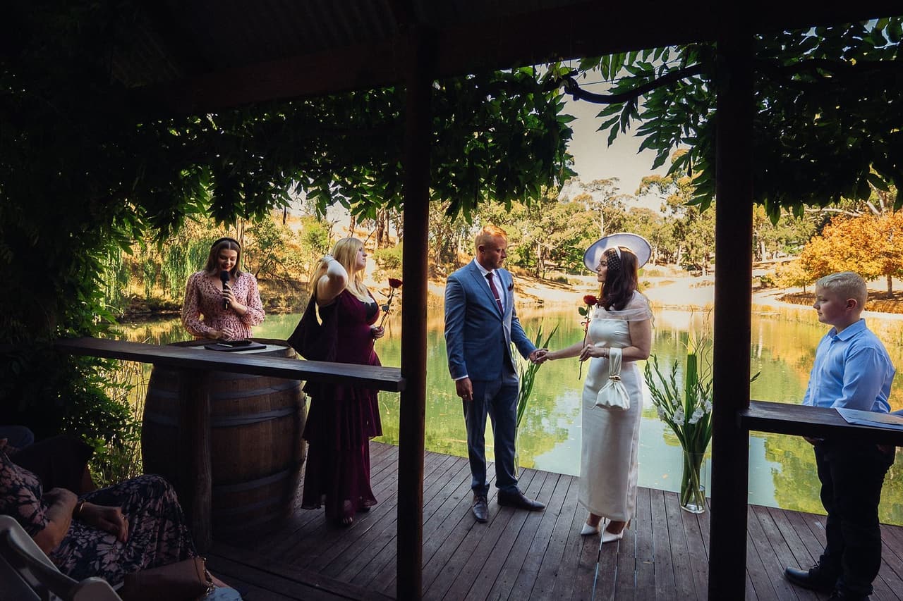 Kim and Ben’s ceremony at the Boatshed, a timber deck under a vine-covered pergola, their guests gathered around