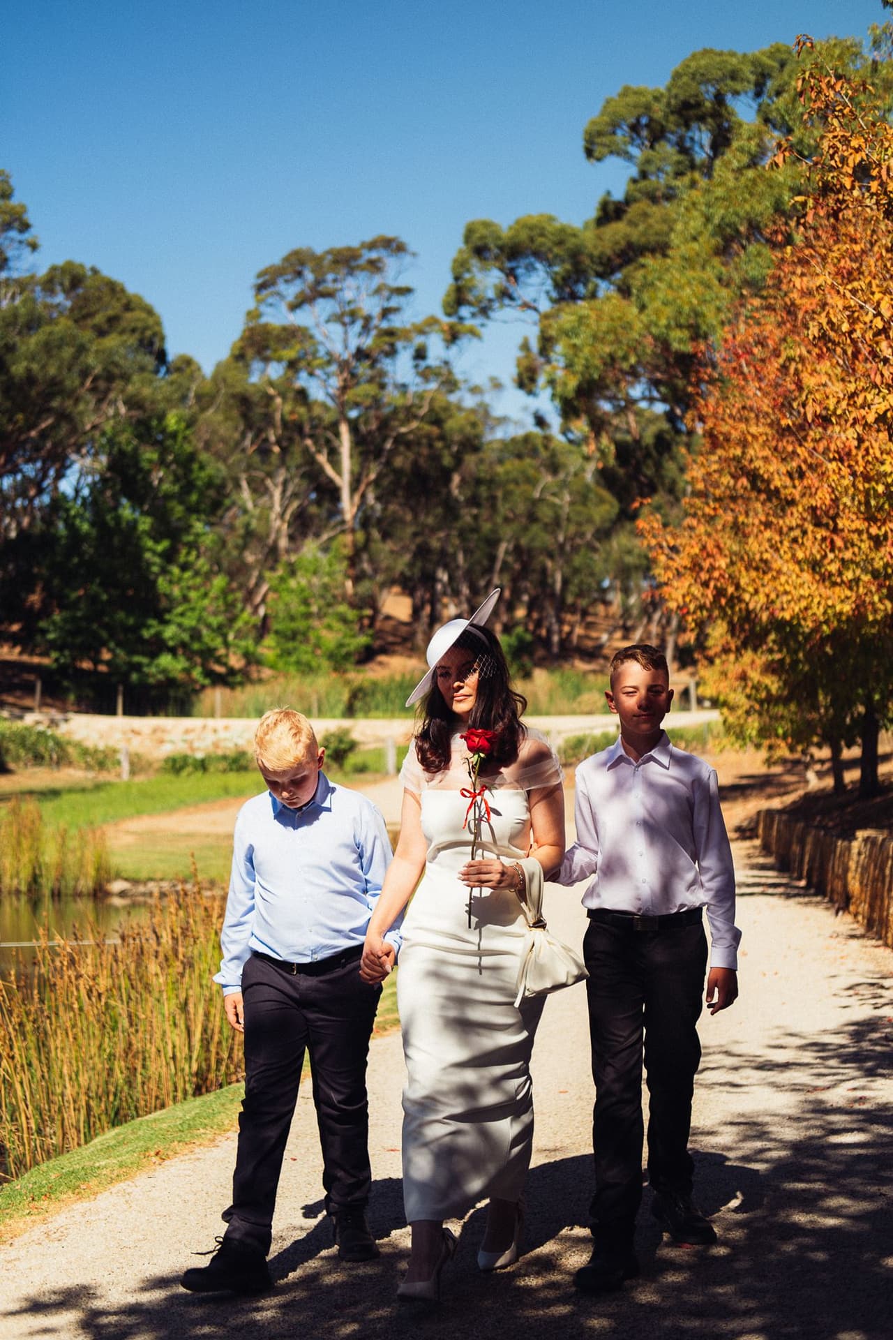 Kim walking down the gravel path with a red rose, a boy on either side of her and autumn trees behind