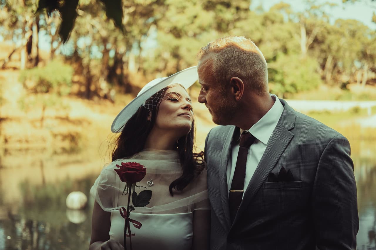 Kim with her eyes closed as Ben leans in close beside the pond, a red rose in her hand
