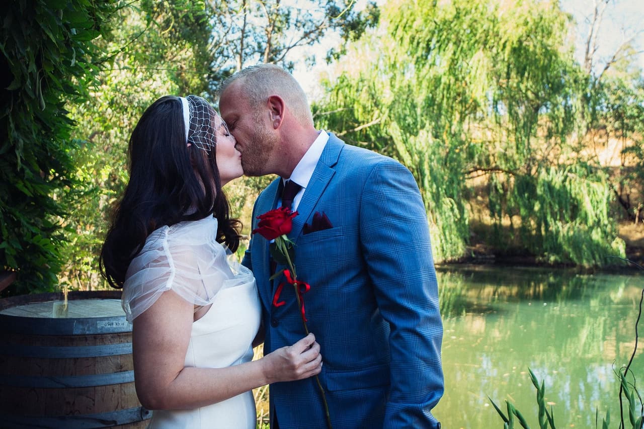 Kim and Ben sharing a kiss beside a wine barrel at the edge of the pond