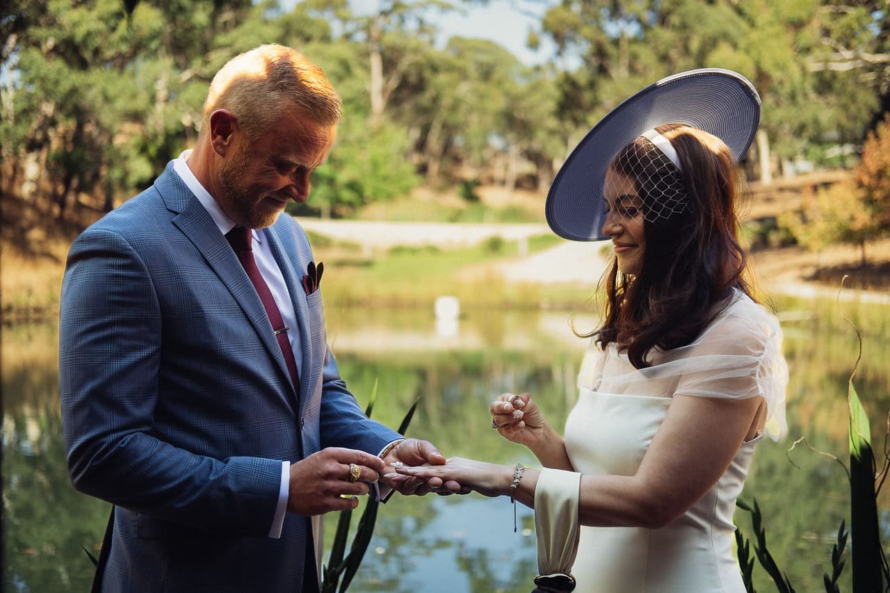 Ben smiling as he slides a ring onto Kim’s finger beside the pond