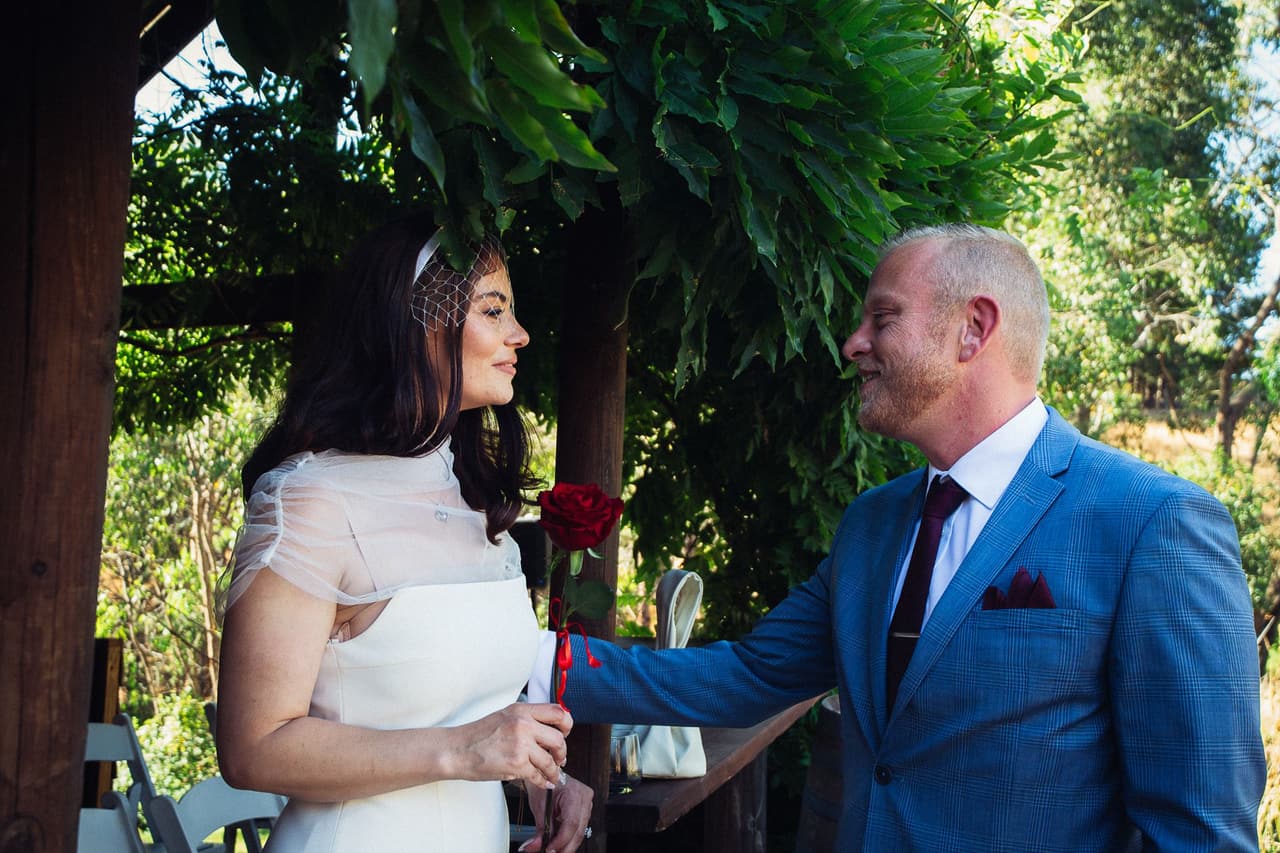 Ben touching the rose in Kim’s hand as they talk under the vines of the Boatshed