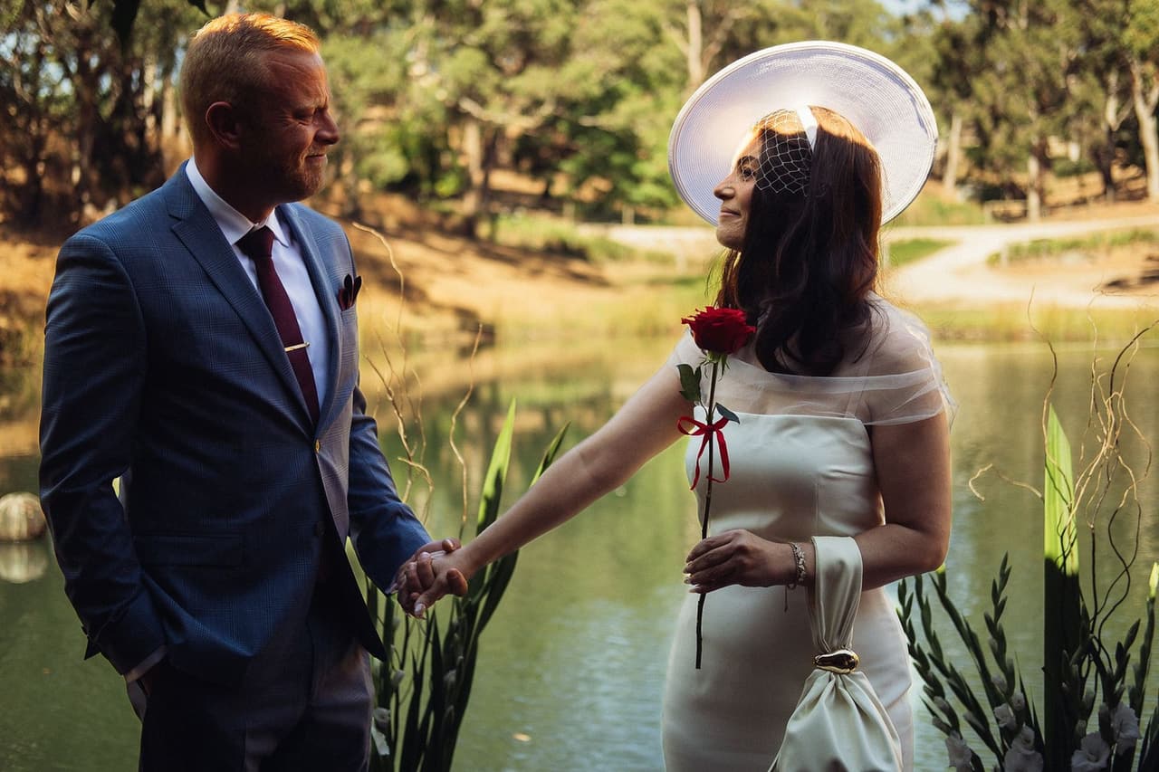 Kim, in a wide-brimmed hat, holding a red rose and Ben’s hand beside the pond