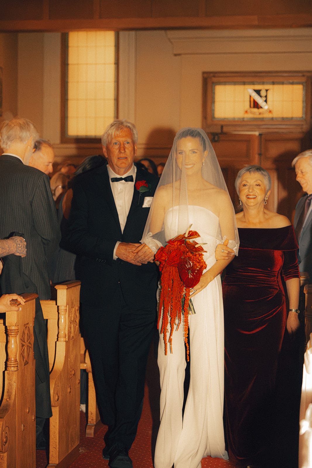 Kira walking down the aisle with two members of her family, red flowers in her arms
