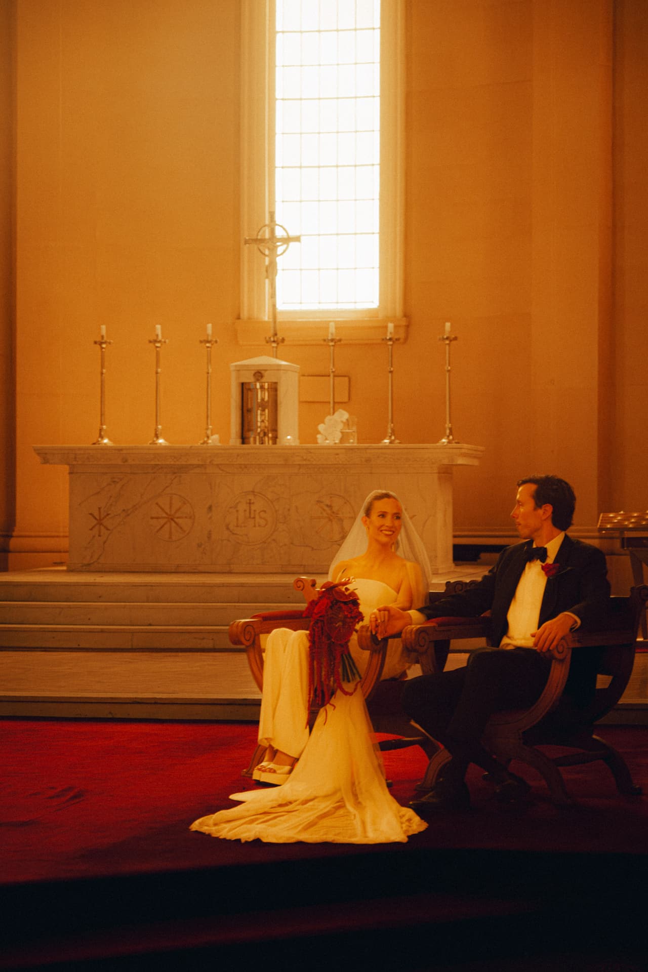 Kira and Nick seated before the altar in warm afternoon light, red roses in her lap