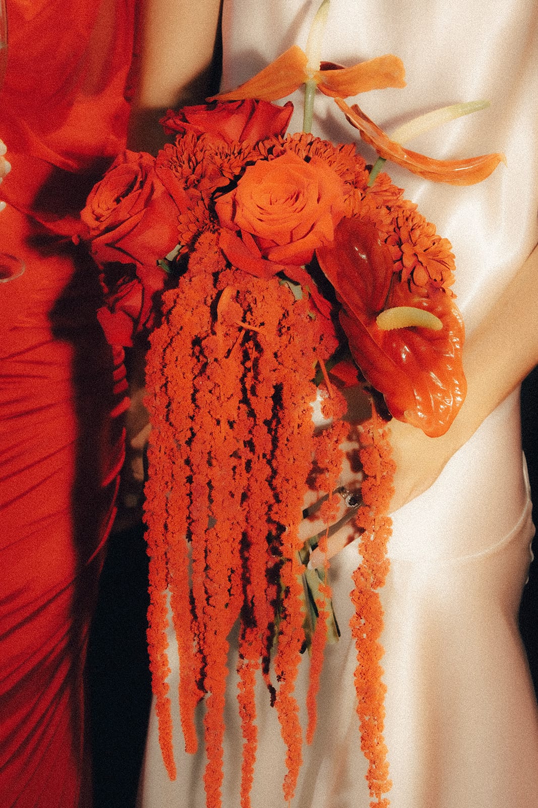Close up of Kira’s cascading bouquet of red and orange roses, anthuriums and amaranthus against her satin dress