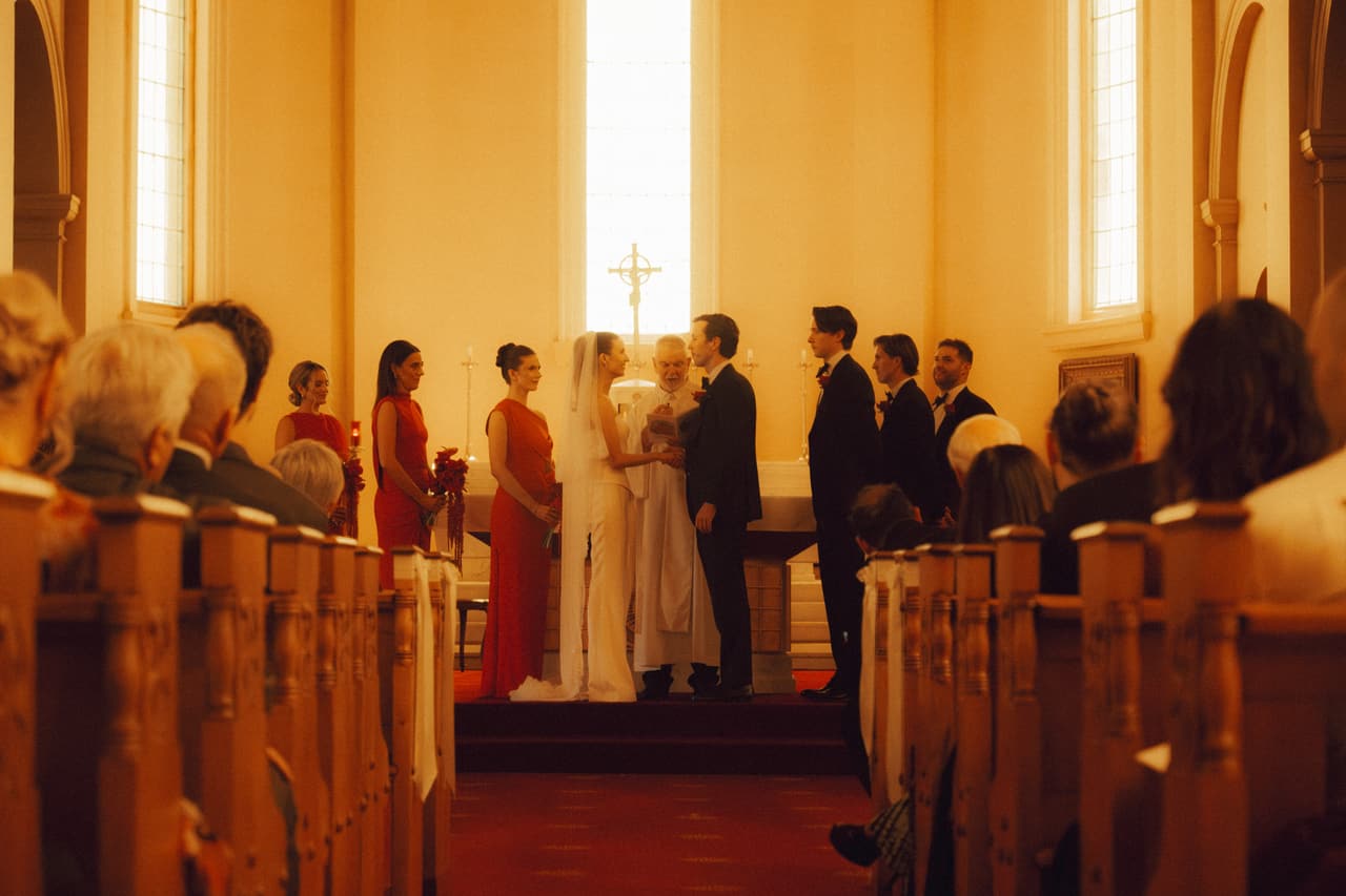 The ceremony seen from the pews, bridesmaids in red beside the altar and sunlight through the high windows