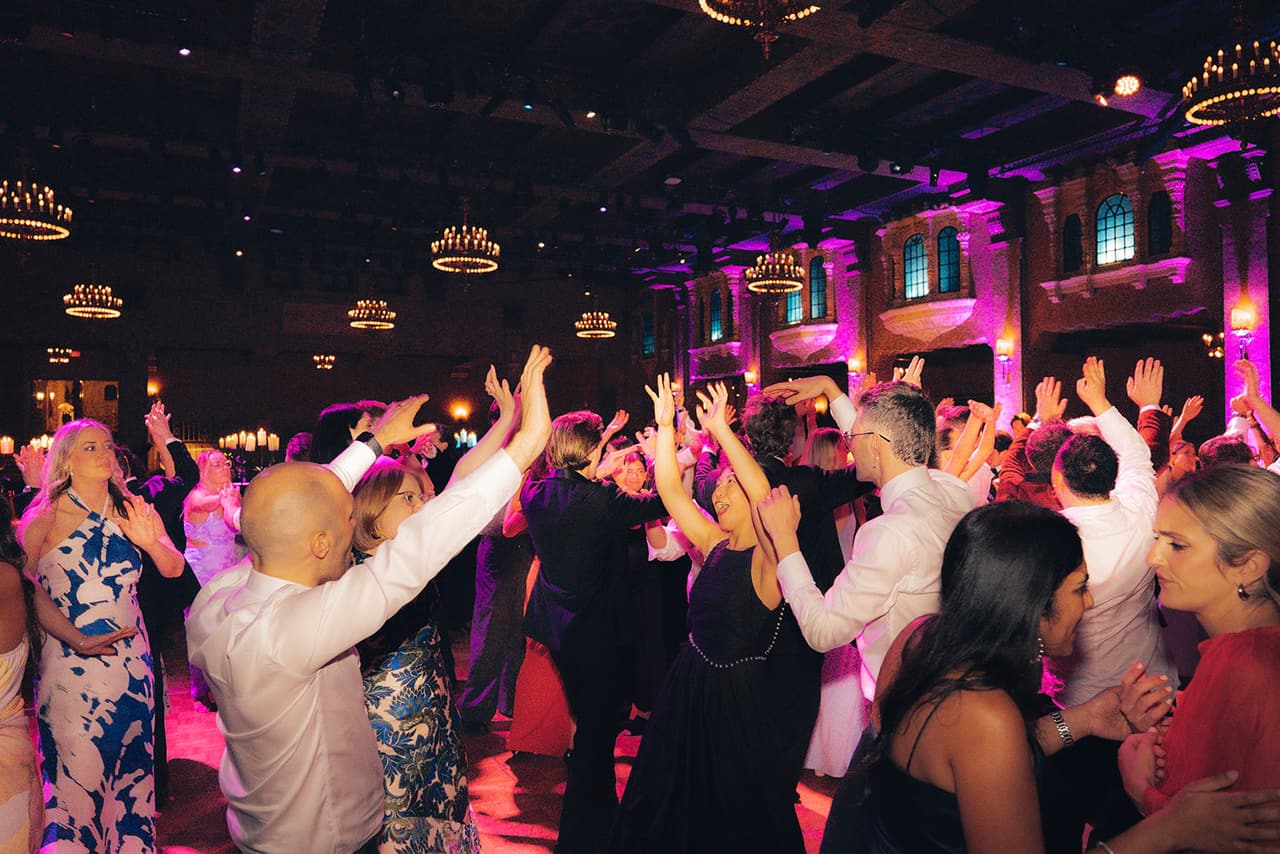Guests with their hands in the air on a packed dance floor under the ballroom’s chandeliers