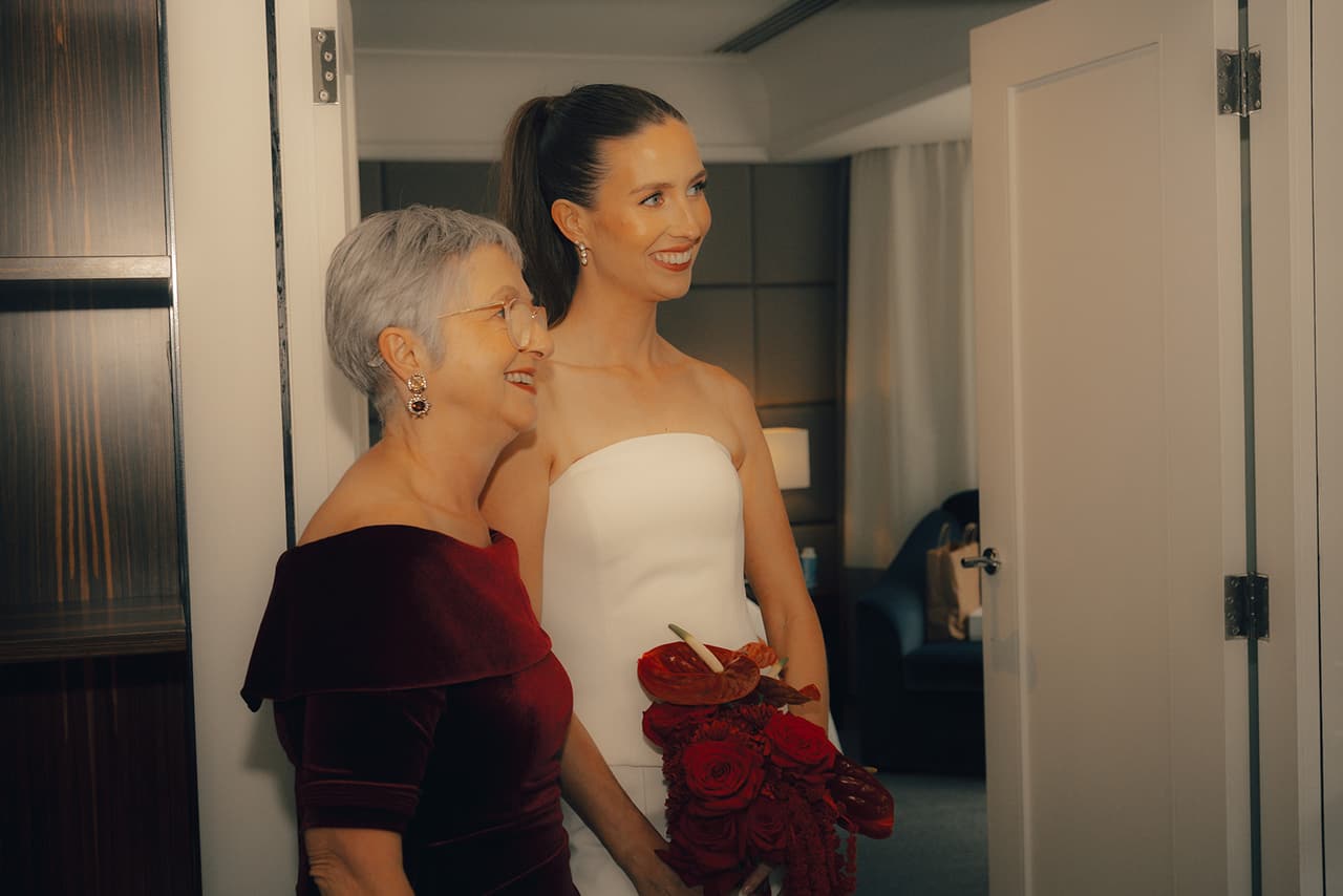 Kira smiling beside an older woman in burgundy velvet in the hotel room, her red bouquet in her hands