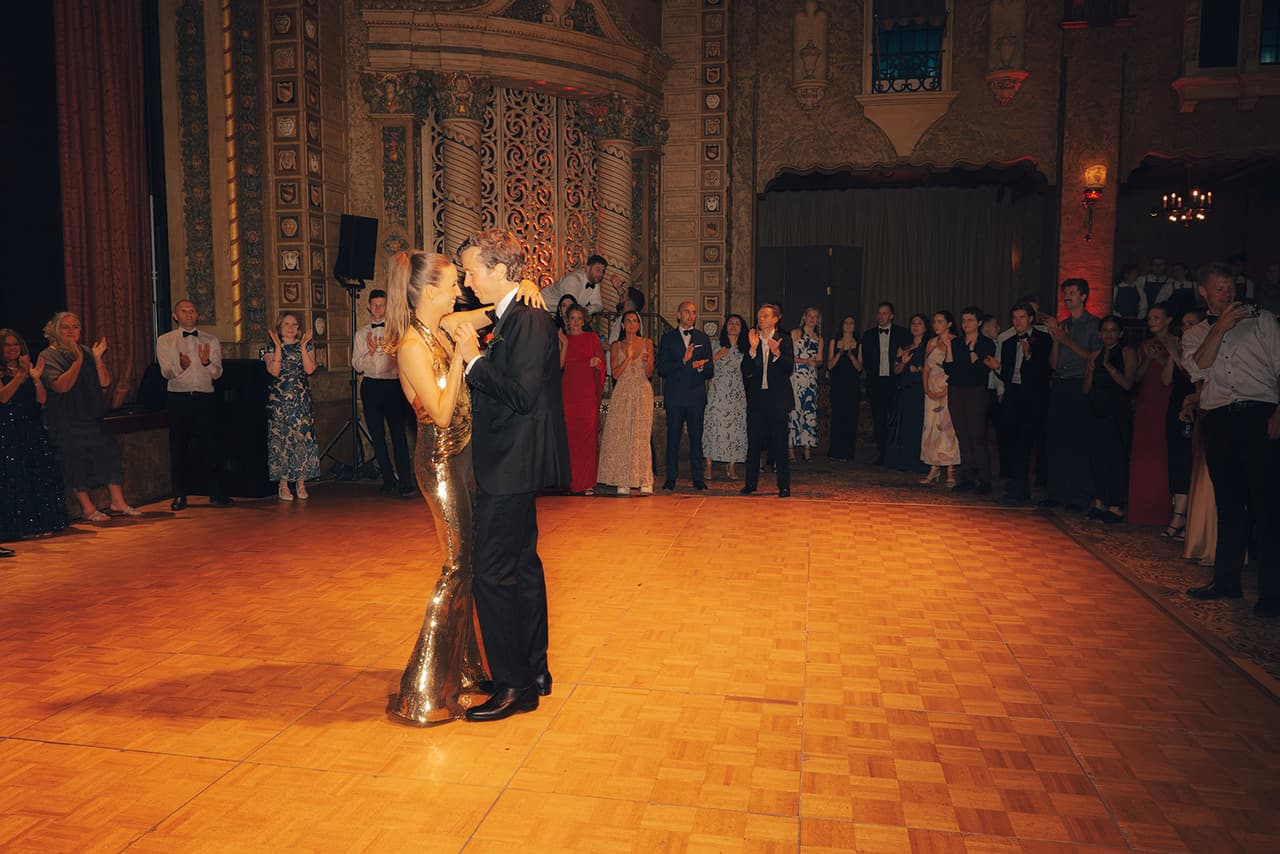 Kira and Nick dancing alone on the parquet floor inside a circle of guests