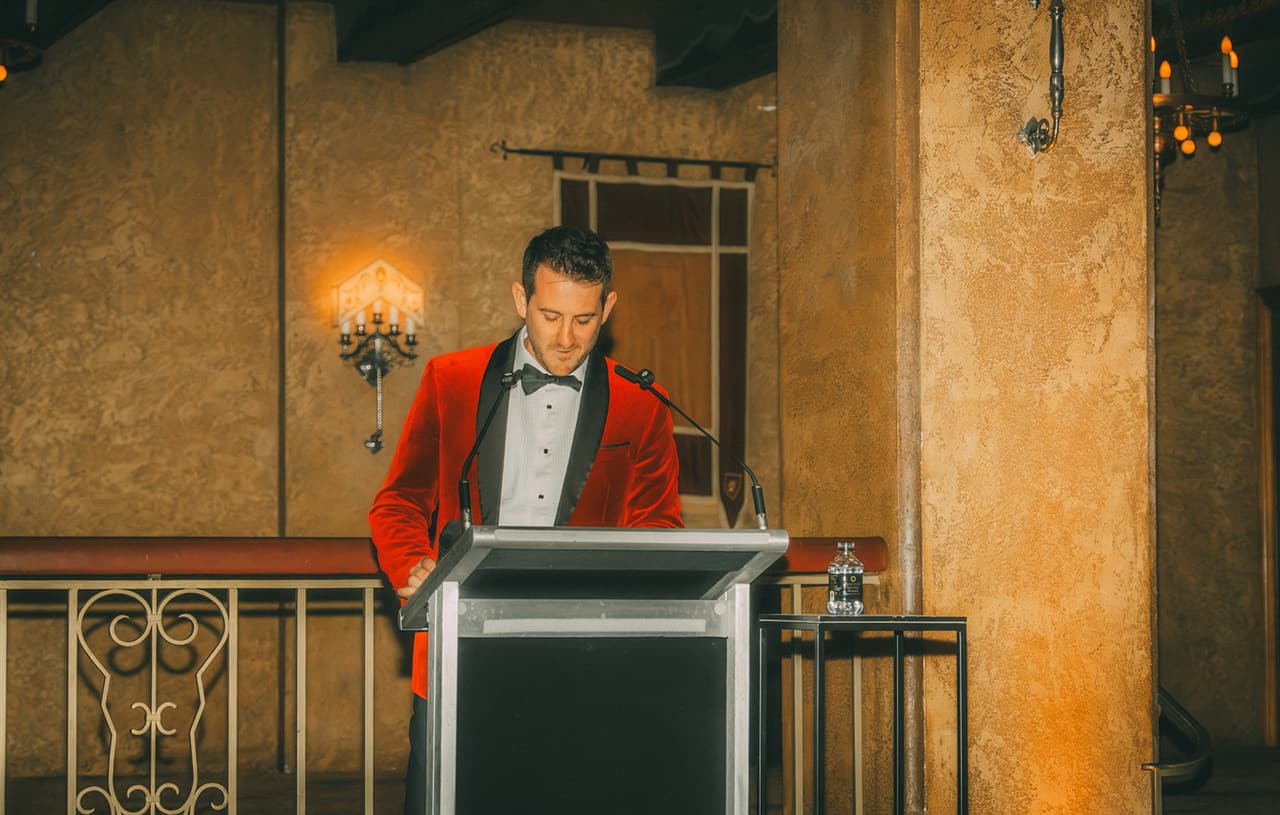 A guest in a red velvet dinner jacket speaking at the lectern in the ballroom