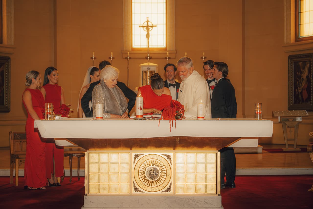 A bridesmaid in red signing the register at the altar beside an older woman, with Kira, Nick and the priest looking on