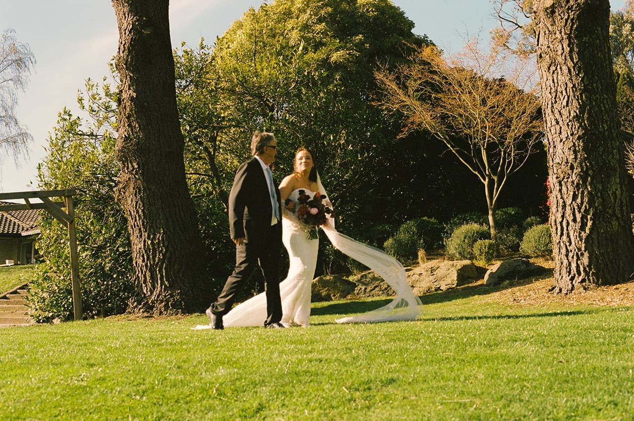 Kristie walking arm in arm across the lawn to the ceremony, her veil trailing behind her, on 35mm film
