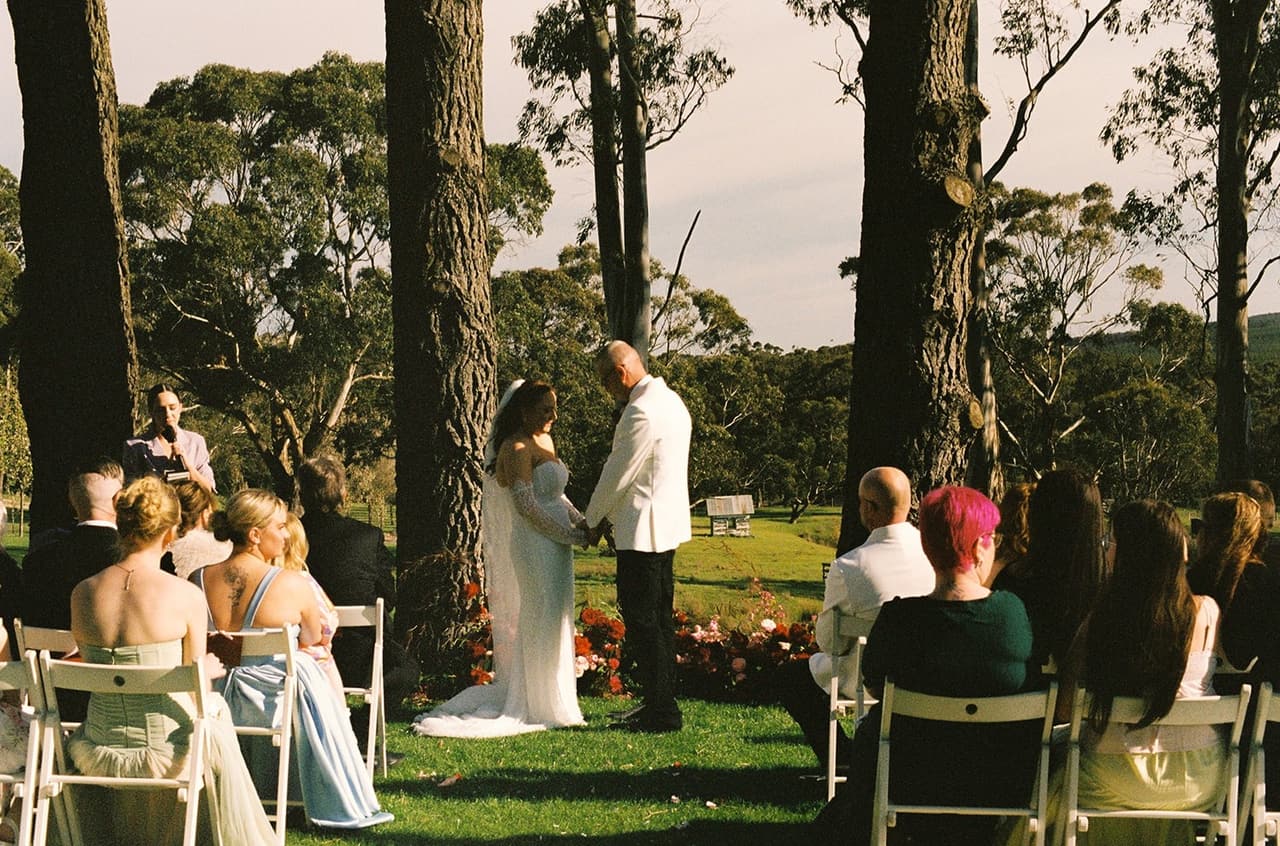 Kristie and Gareth holding hands at the front of the ceremony under the tall trees, seen down the aisle between their guests, on 35mm film