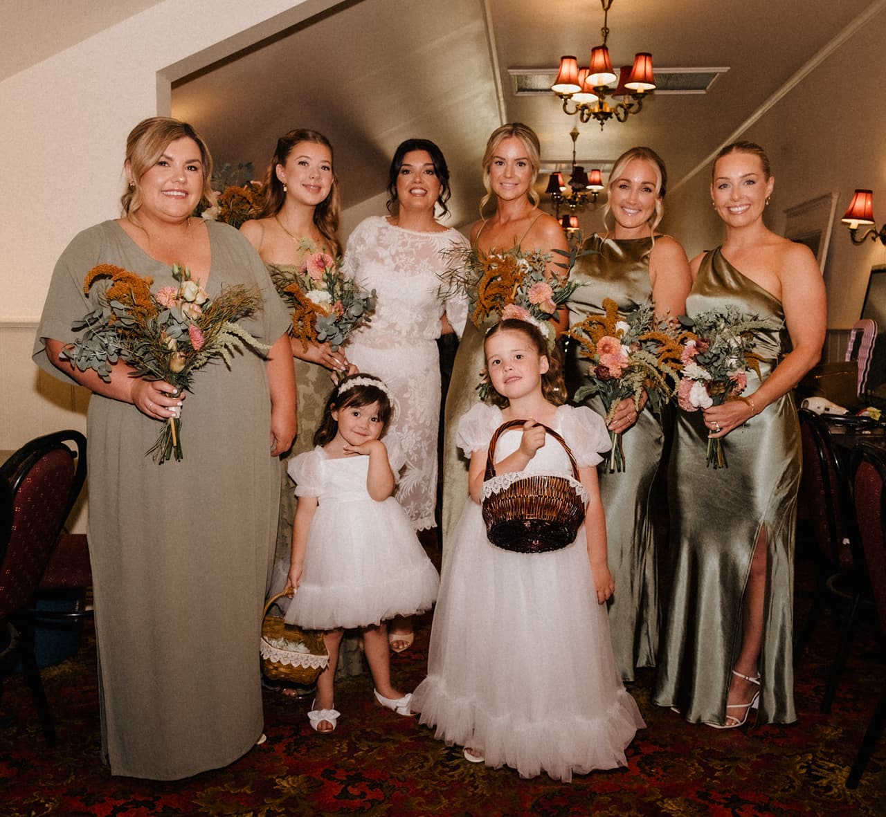 Lana in white lace with her bridesmaids in sage green and two flower girls, under the chandeliers of the room where they got ready