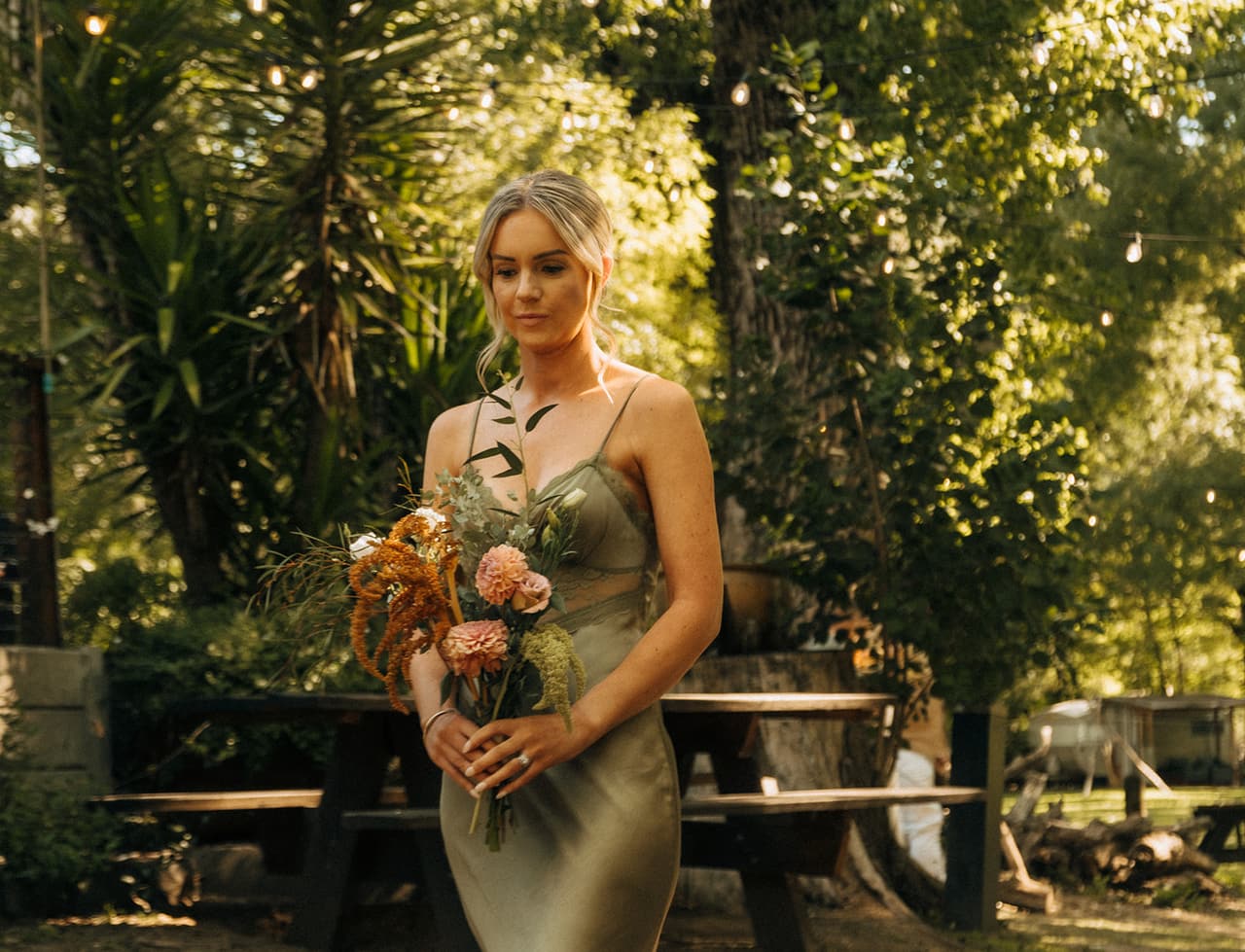 A bridesmaid in sage green walking through the trees with her bouquet