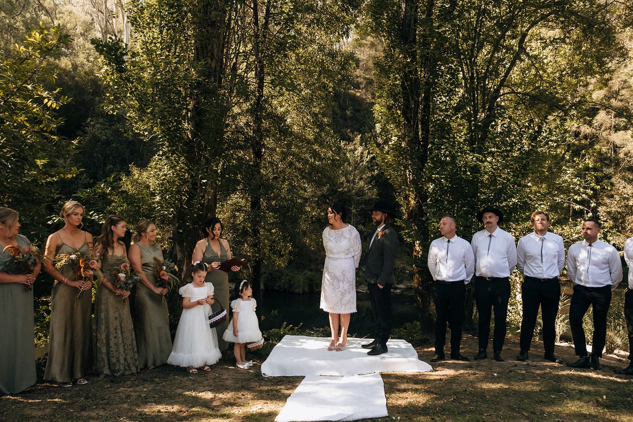 The ceremony in a shady grove, bridesmaids in sage green on one side and groomsmen in white shirts on the other