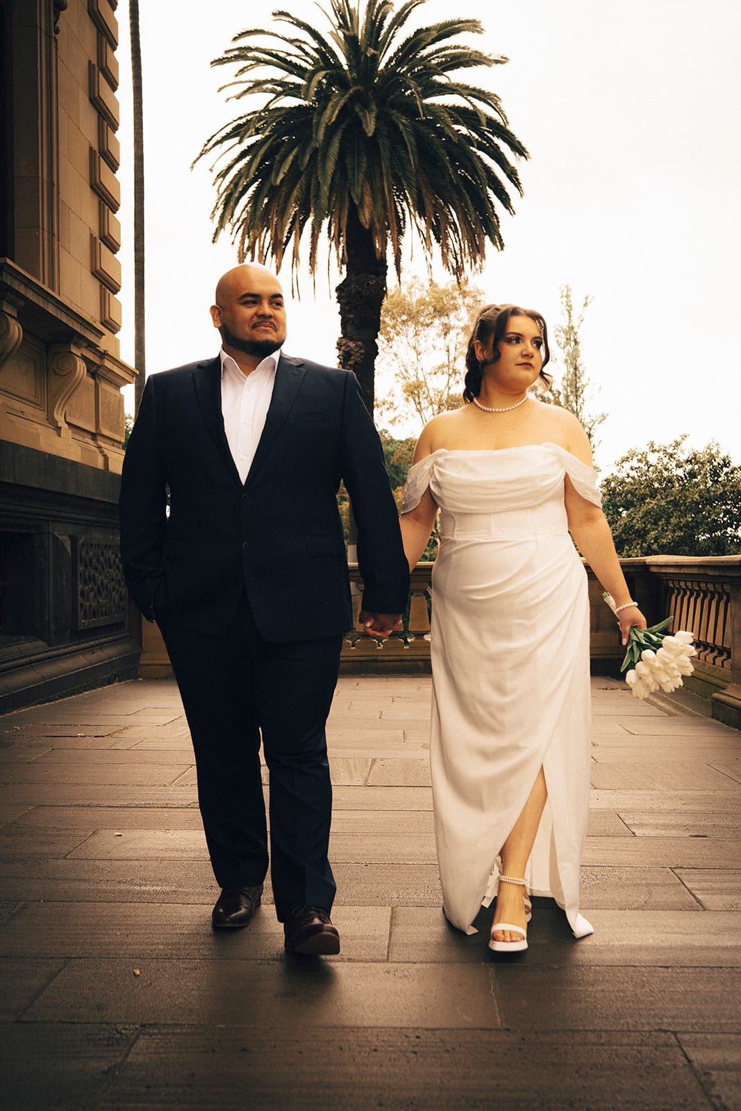 Martina and Diego walking hand in hand along the balcony, a palm tree behind them
