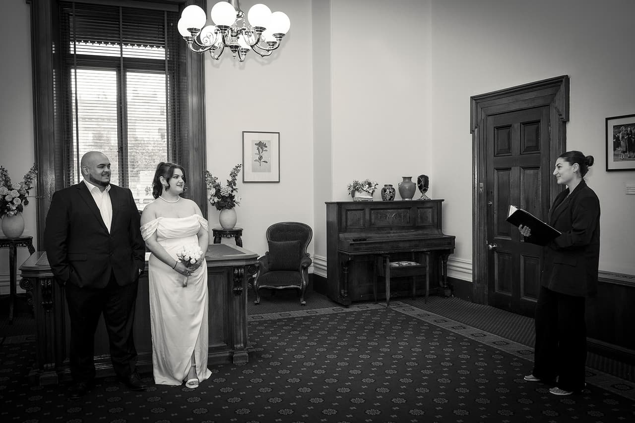 Black and white photo of the celebrant reading to Martina and Diego across the ceremony room, a piano against the wall